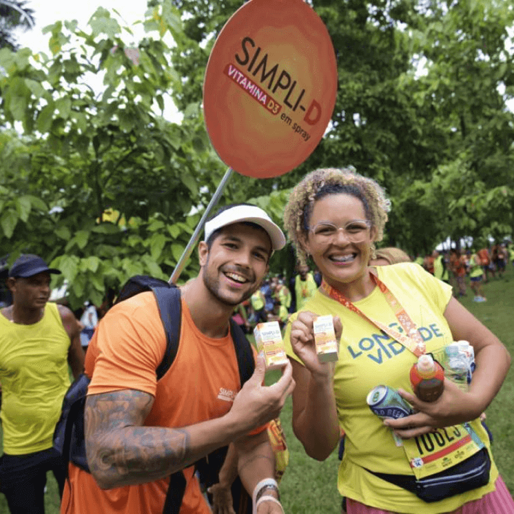 Duas pessoas sorrindo, segurando caixas do Simpli-D na corrida do evento. Homem carregando a placa de divulgação da vitamina.