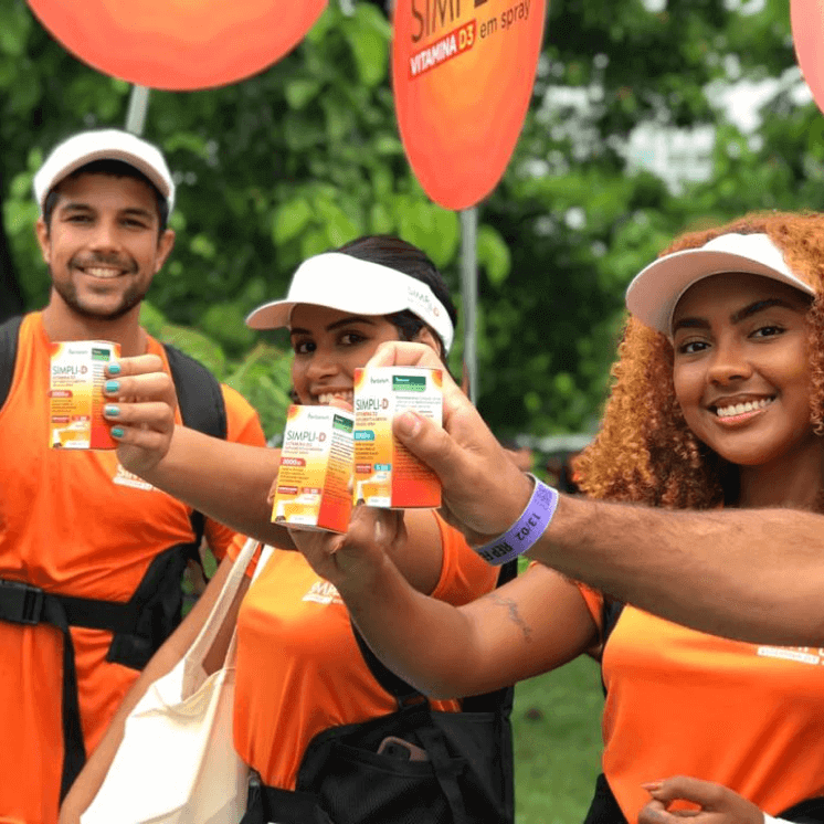 Três pessoas sorrindo, segurando caixas do Simpli-D, divulgando as vitaminas.